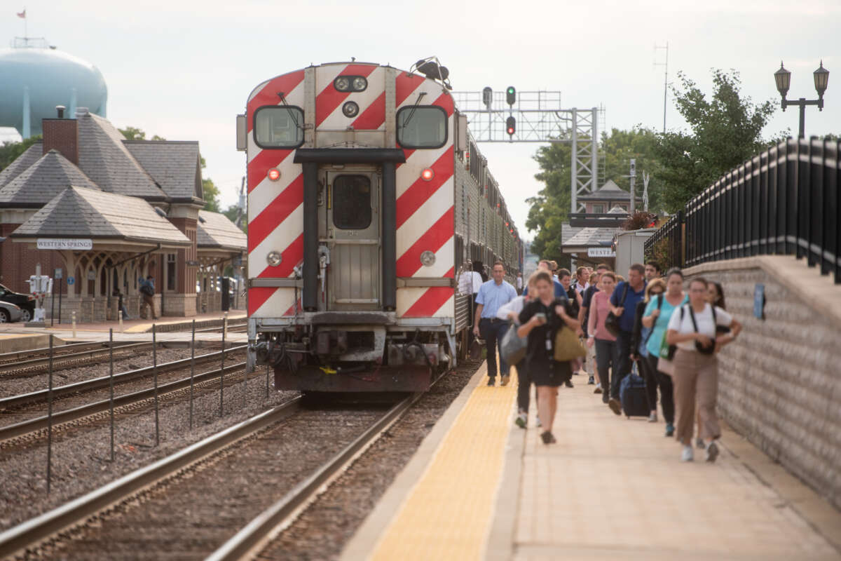 Group of people walking on sidewalk alongside a Metra train stopped on tracks.
