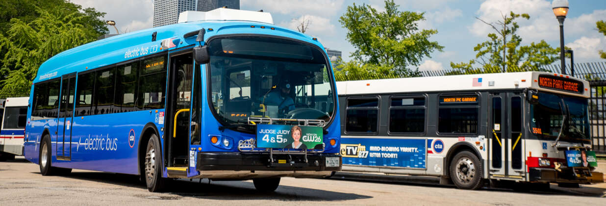An electric CTA bus and a diesel CTA bus with skyscrapers and trees in the background.