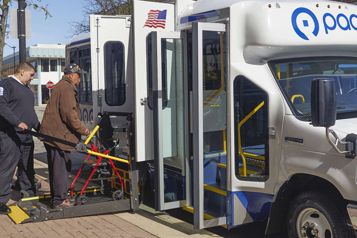 A Pace Paratransit driver assists a man with a walker in getting into the vehicle using a lift.